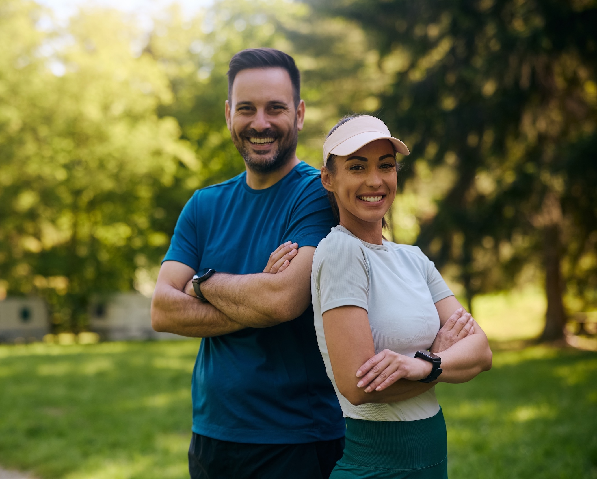 A smiling couple in athletic wear standing back to back with their arms crossed in a sunlit park